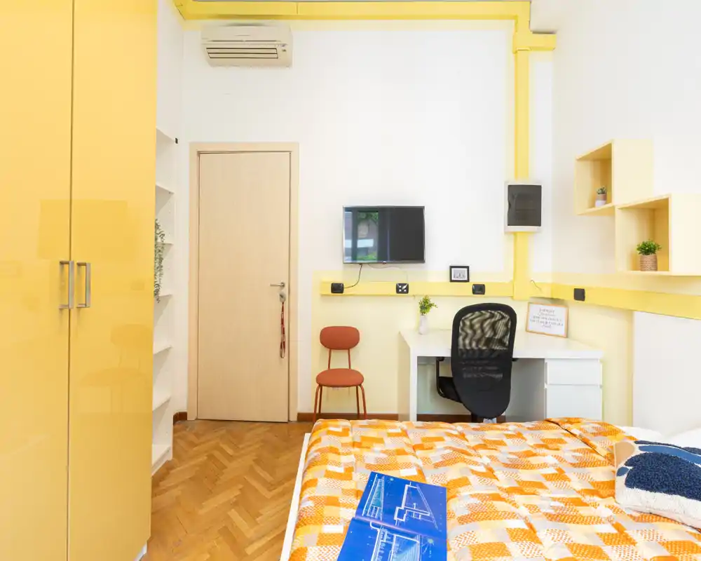 Dormitorio luminoso y compacto con colcha naranja estampada, escritorio integrado, estanterías, armario y pequeña TV — ideal para estudiante o una persona sola.