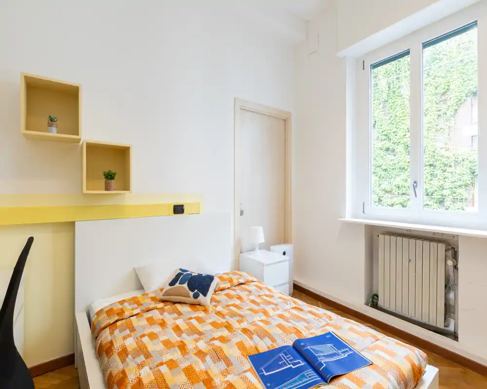 Dormitorio luminoso y ordenado con cama individual de ropa de cama naranja, mesita con lámpara y una gran ventana que aporta abundante luz natural.