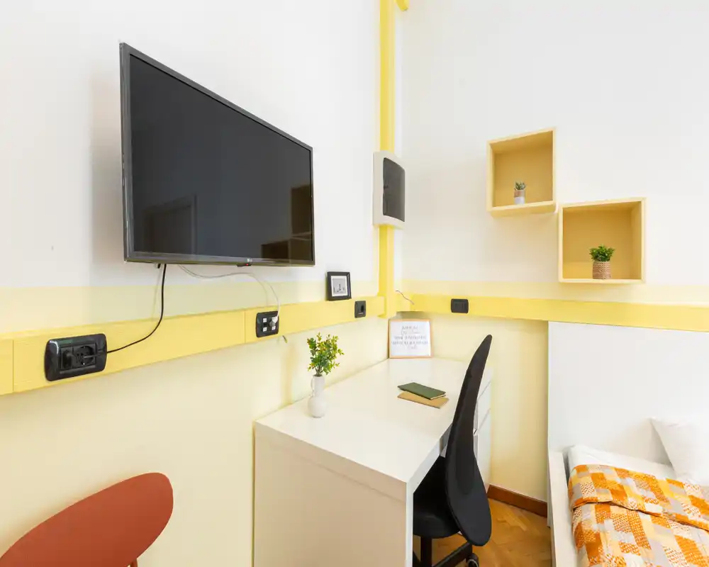 Rincón de dormitorio luminoso y compacto con escritorio pequeño, televisor en la pared y una cama individual parcialmente visible con manta naranja y estantes decorativos.