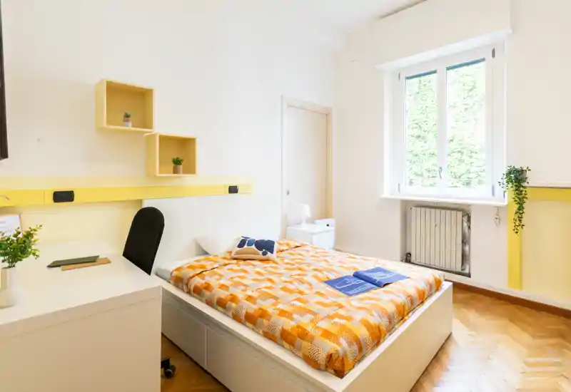 Habitación individual moderna y luminosa con cama central vestida con una cálida colcha naranja, zona de escritorio y abundante luz natural por una gran ventana.