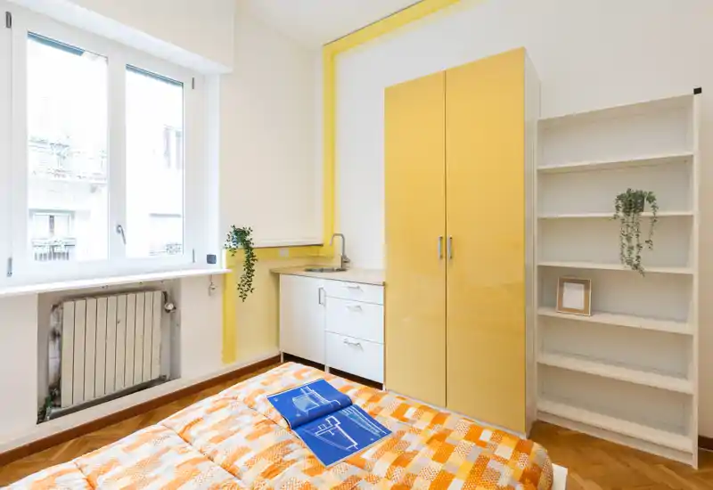 Dormitorio compacto y luminoso con colcha naranja estampada, armario integrado y una pequeña zona con lavabo. Ventana amplia que aporta luz natural y decoración ordenada con plantas y estanterías.
