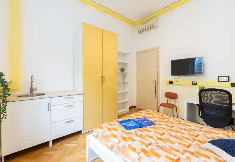 Habitación individual luminosa y bien preparada con colcha naranja estampada, armario amarillo, pequeño fregadero de cocina y escritorio con TV — espacio compacto y alegre, ideal para estudiantes o estancias cortas.