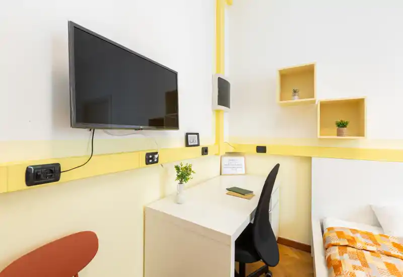 Rincón de dormitorio luminoso y compacto con escritorio pequeño, televisor en la pared y una cama individual parcialmente visible con manta naranja y estantes decorativos.