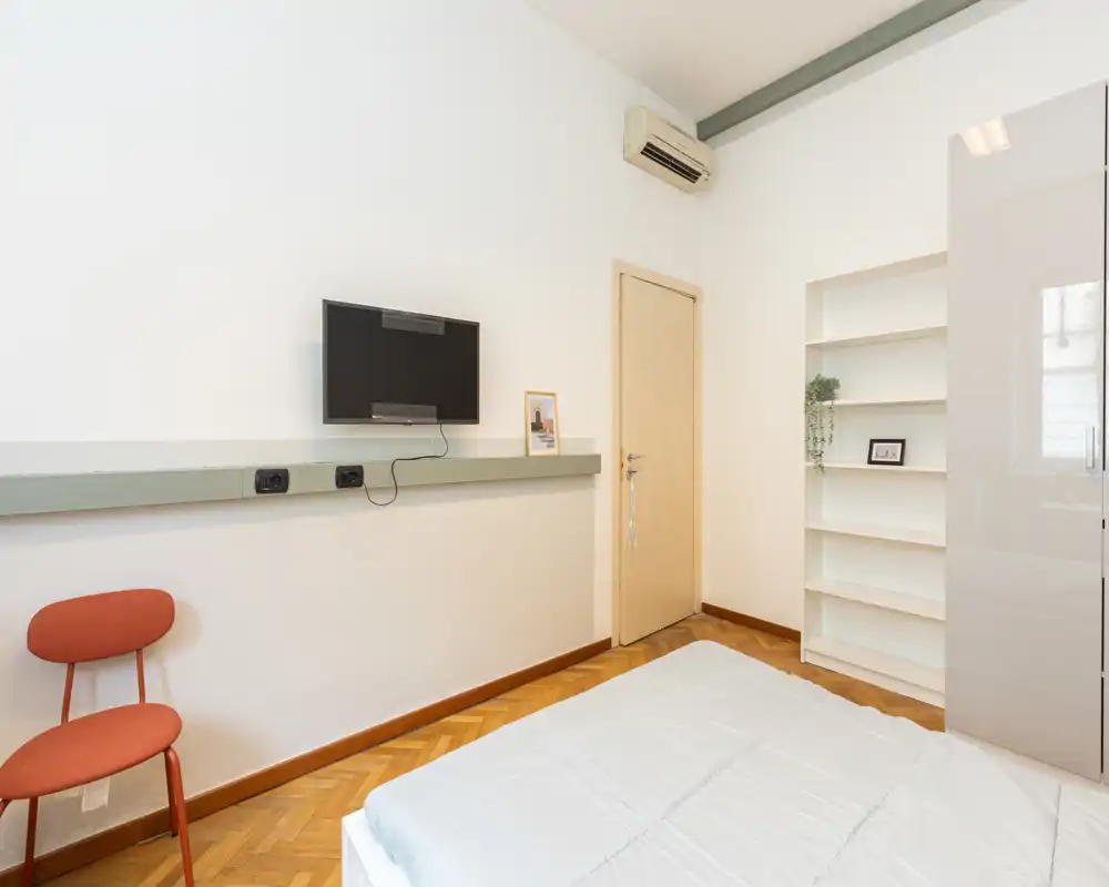 Dormitorio luminoso y minimalista con cama doble bien tendida, televisor en pared y estanterías empotradas — líneas modernas y luz natural que lo hacen acogedor.