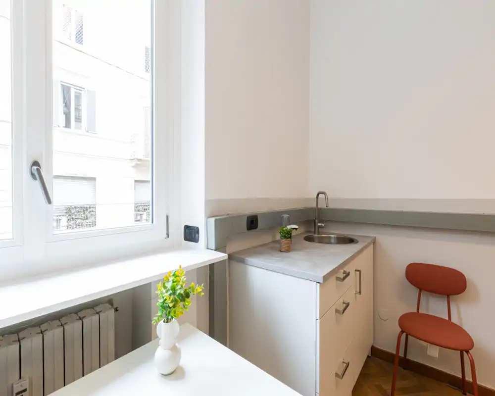Kitchenette compacta y moderna con fregadero de acero, encimera clara y una silla acogedora junto a una ventana luminosa — ideal para un estudio.