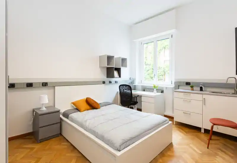 Habitación individual luminosa y moderna con cama bien arreglada, zona de estudio junto a la ventana y pequeño rincón de cocina. Líneas limpias y luz natural crean un ambiente cómodo y ventilado, perfecto para un anuncio de alquiler.