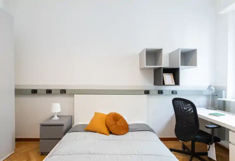 Habitación individual moderna y luminosa con cama bien hecha, dos cojines decorativos, mesita con lámpara y escritorio con silla junto a la ventana — diseño limpio y minimalista, ideal para estudiantes o profesionales.