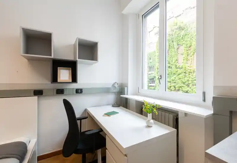 Rincón de dormitorio luminoso y compacto con escritorio blanco junto a una gran ventana, silla ergonómica y una pequeña porción de la cama visible. Luz natural y estanterías minimalistas para un espacio de trabajo funcional.