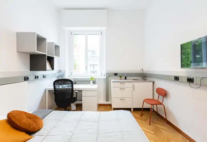 Dormitorio luminoso y compacto con cama bien arreglada, escritorio junto a la ventana y pequeño rincón con fregadero — ideal para una persona o estudiante.