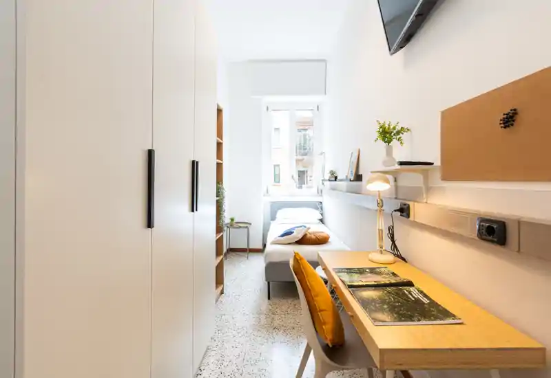 Dormitorio individual luminoso con escritorio moderno y amplio armario. La luz natural, cojines acogedores y una lámpara de lectura crean un espacio ideal para estudiar y descansar.