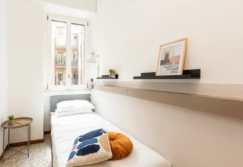 Dormitorio individual luminoso y compacto con cama bien tendida bajo la ventana, cojines decorativos y estanterías minimalistas—ideal para estancias cortas y cómodas.