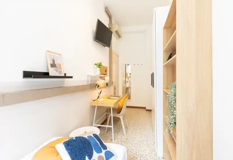 Rincón de dormitorio luminoso y compacto con una pequeña cama visible, un escritorio estrecho y TV de pared — ideal para una persona. El espacio está decorado de forma minimalista con tonos de madera cálidos y cojines que aportan confort.