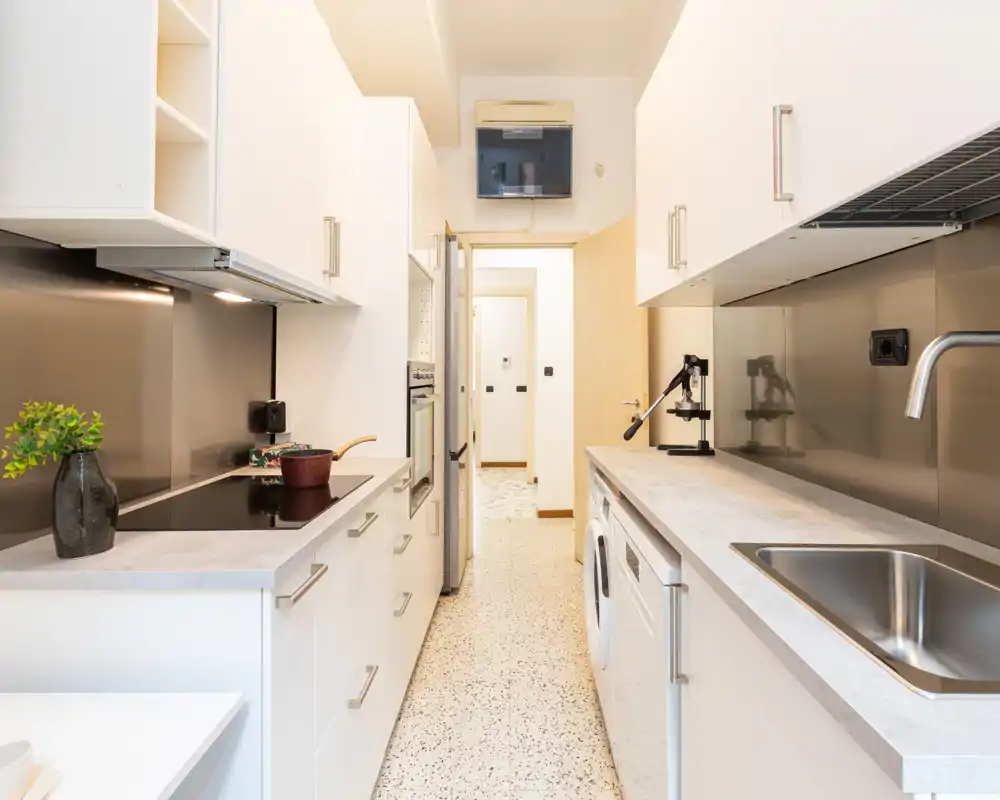 Cocina moderna tipo pasillo, luminosa, con armarios blancos, salpicadero metálico, fregadero integrado, placa de inducción y electrodomésticos empotrados — ordenada y lista para usar.