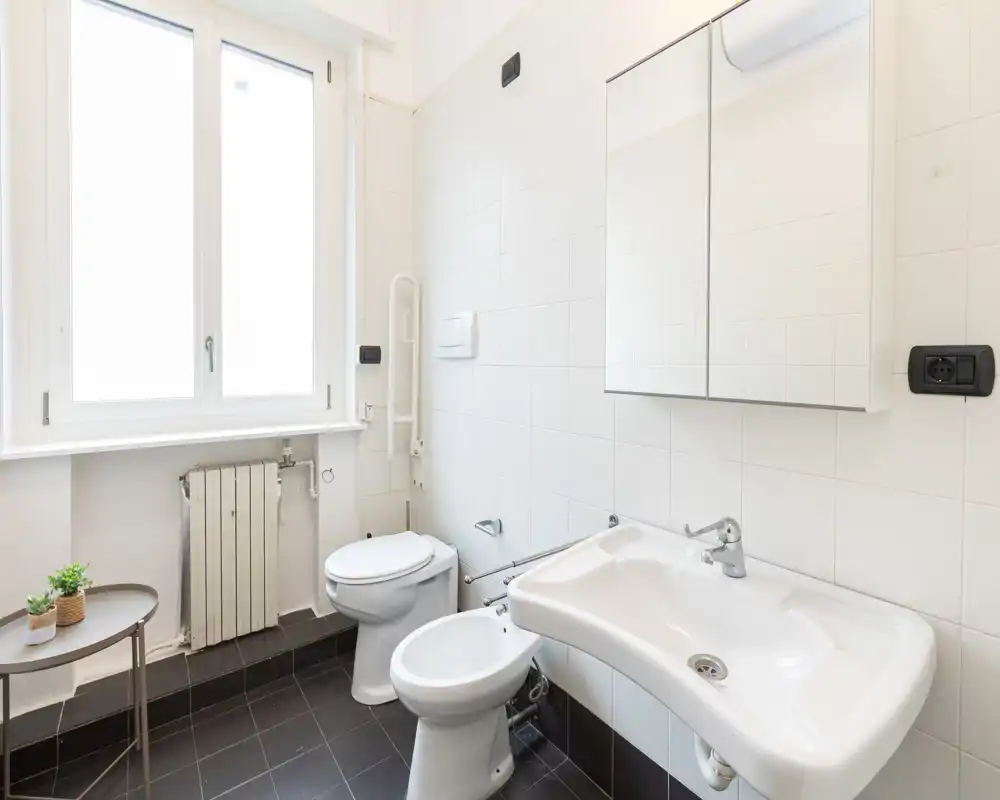 Baño luminoso y limpio con lavabo, bidé, inodoro y armario con espejo; ventana grande que aporta luz natural y suelo de baldosas oscuras.