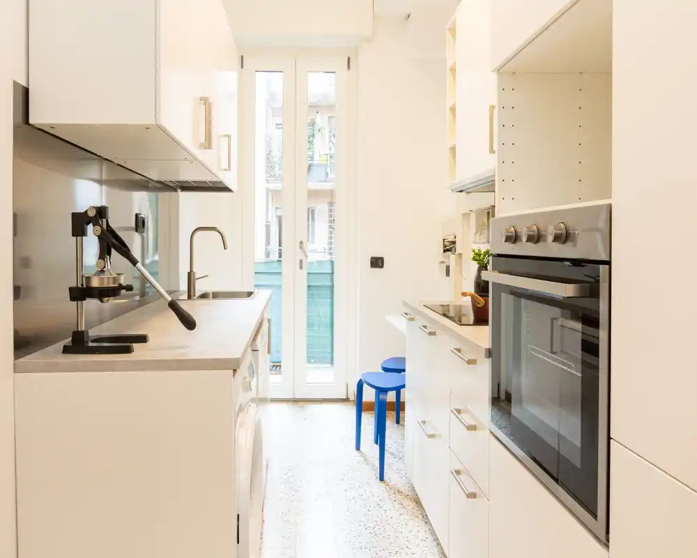 Cocina moderna tipo pasillo, luminosa, con muebles blancos, horno empotrado, pequeños electrodomésticos sobre la encimera y puerta de cristal que aporta luz natural.