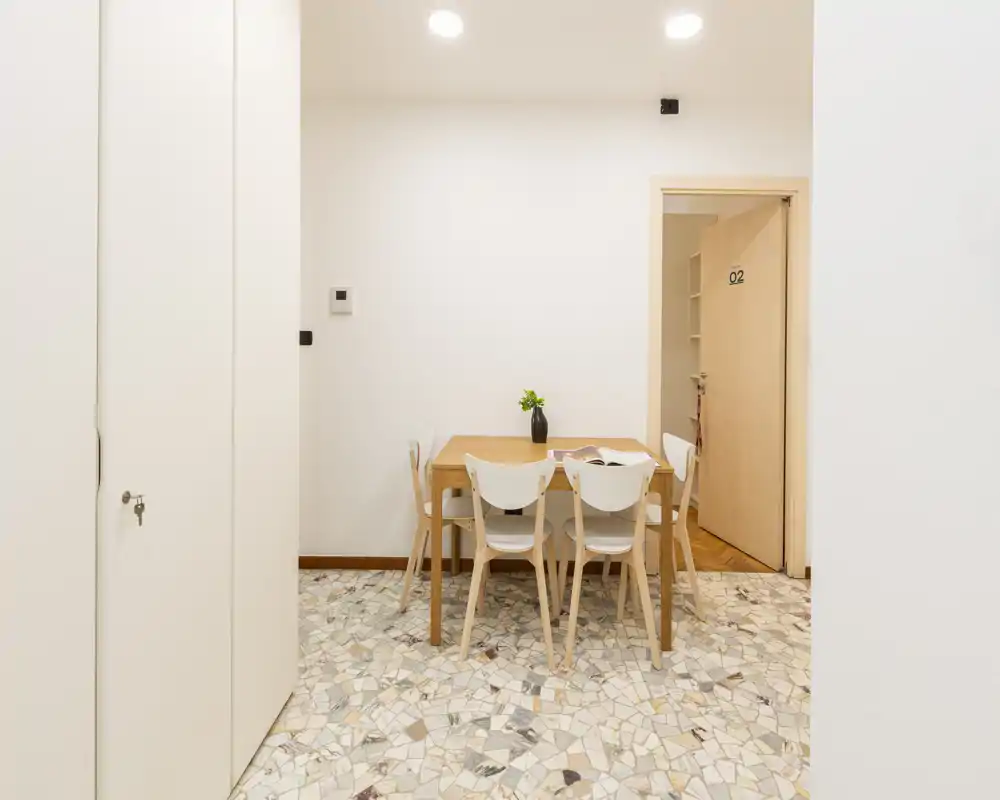 Zona de comedor minimalista con mesa de madera compacta y cuatro sillas modernas, paredes neutrales luminosas y suelo de terrazo; decorada con un pequeño jarrón.