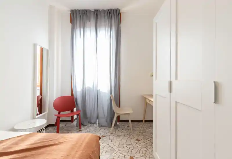 Dormitorio luminoso y minimalista con tonos cálidos en la cama, armario y luz natural a través de una cortina translúcida. Suelo de mosaico, dos sillas y una pequeña mesa que hacen la habitación acogedora para una o dos personas.