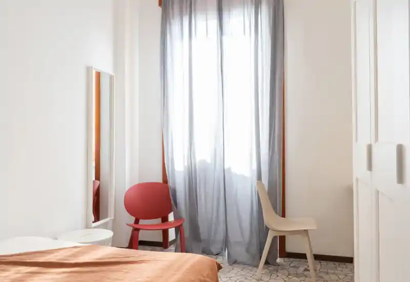 Rincón de dormitorio luminoso y minimalista con cama parcialmente visible, cortinas translúcidas que dejan pasar luz natural y dos sillas sencillas que crean un ambiente sereno.