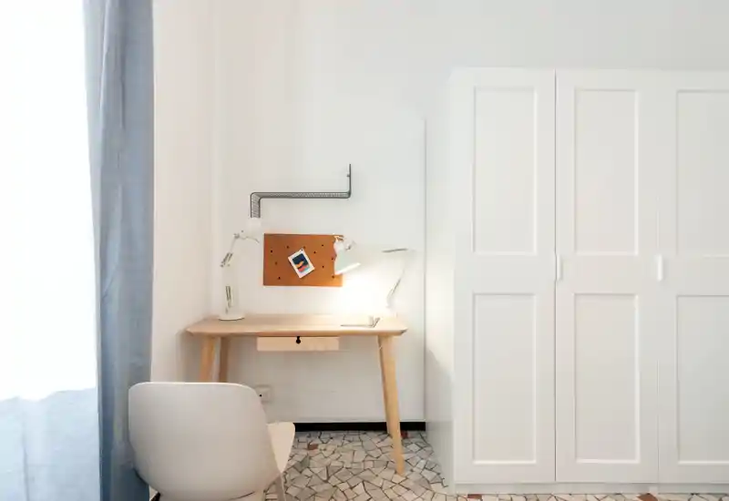 Rincón de dormitorio minimal y luminoso con un escritorio de madera, una silla y un armario blanco junto a una ventana que aporta luz natural.