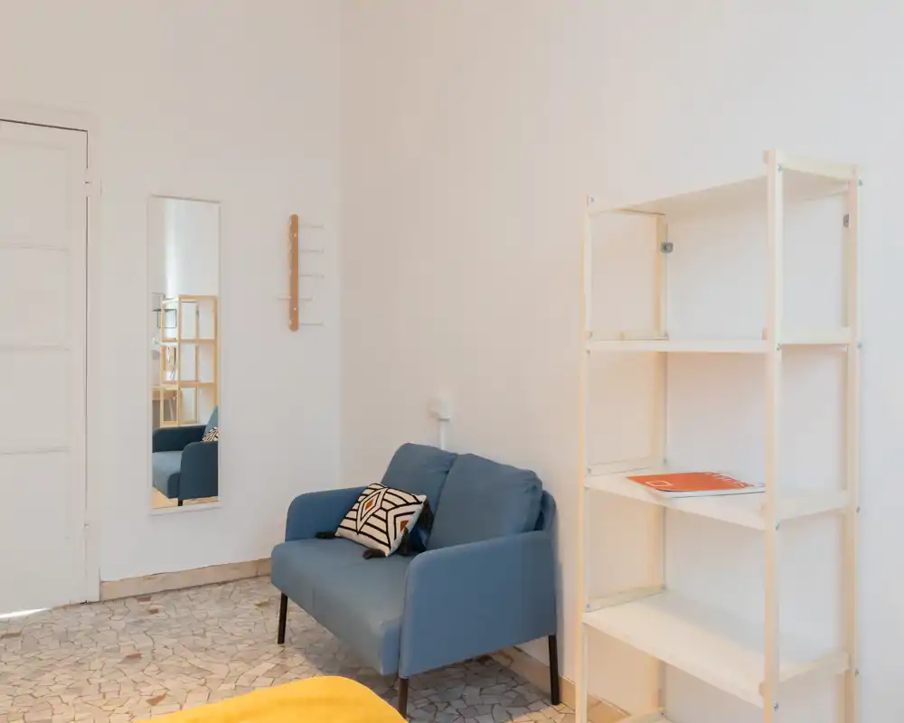 Small, minimal living area with a two-seater blue sofa, decorative cushion, full-length mirror and open shelving — bright, clean and simply styled.