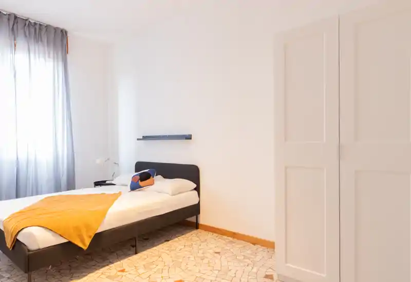 Dormitorio luminoso y minimalista con cama doble, manta cálida y una lámpara de mesita; la luz natural de una ventana alta crea un ambiente sereno.