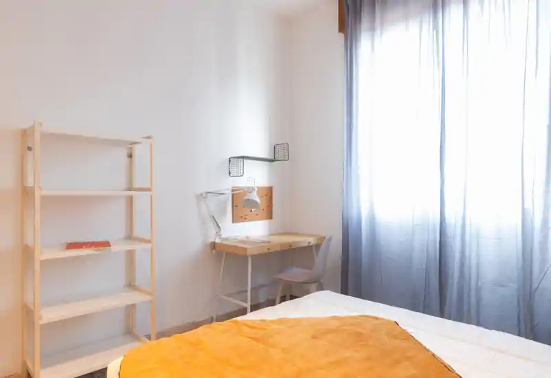 Dormitorio luminoso y minimalista con cama bien hecha, escritorio sencillo y estantería junto a una gran ventana que deja entrar luz natural suave.