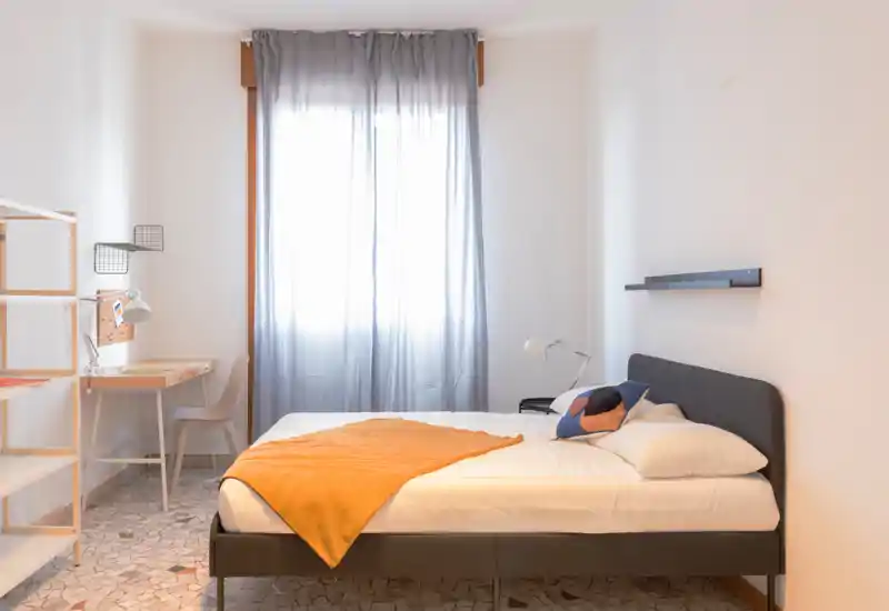 Dormitorio luminoso y minimalista con cama doble, lámpara de noche y un pequeño escritorio junto a la ventana; textiles suaves y una manta cálida.