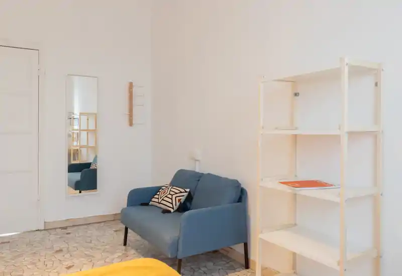 Pequeña sala de estar minimalista con sofá azul de dos plazas, cojín decorativo, espejo de cuerpo entero y estantería abierta — luminosa, limpia y de estilo sencillo.