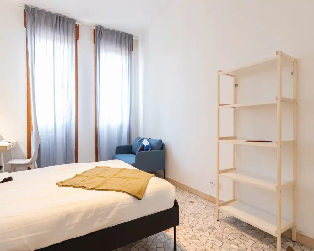 Dormitorio luminoso y minimalista con una cama grande, luz natural de ventanas altas, un sillón acogedor y estantería sencilla — ideal para una estancia tranquila.