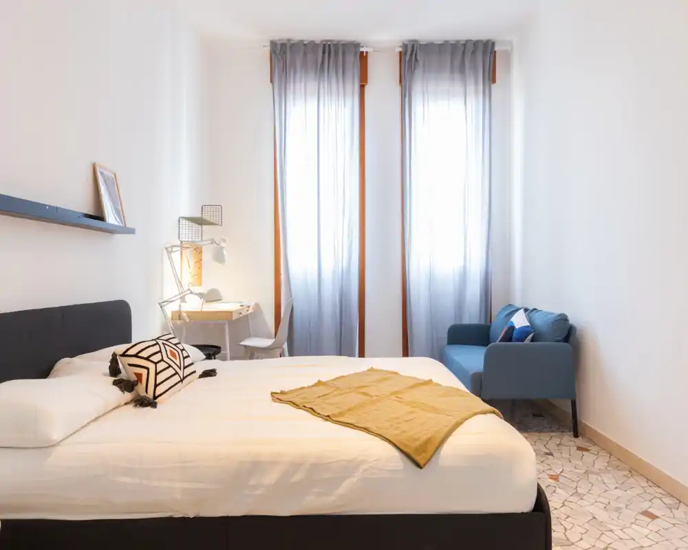 Dormitorio moderno y luminoso con una cama grande, escritorio pequeño y un sofá azul junto a ventanas altas que dejan entrar luz natural.