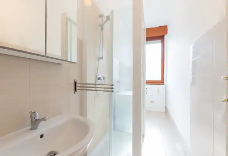 Baño luminoso y compacto con lavabo, armario con espejo y ducha con mampara de cristal. Azulejos blancos y luz natural desde la ventana que aportan sensación de amplitud.