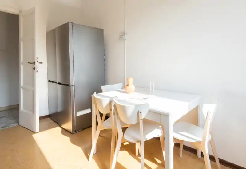 Rincón de cocina minimalista y luminoso con mesa para cuatro, frigorífico moderno y luz natural — ideal para estancias urbanas compactas.