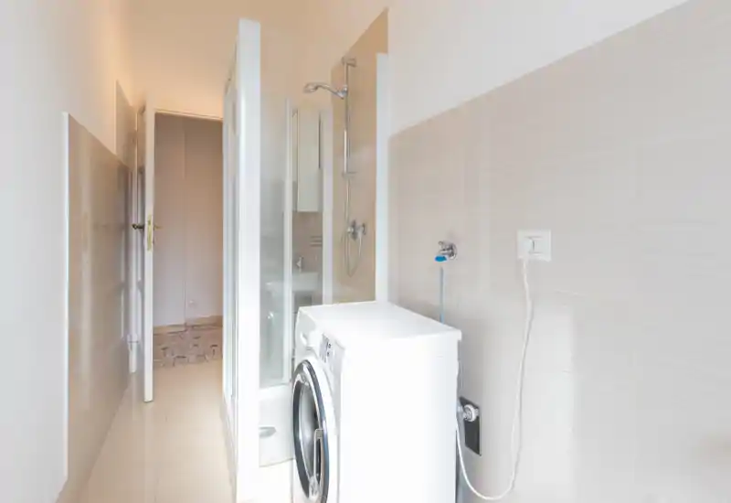 Baño compacto con ducha en esquina y lavadora de carga frontal; azulejos claros y grifería moderna que muestran un espacio funcional y limpio.