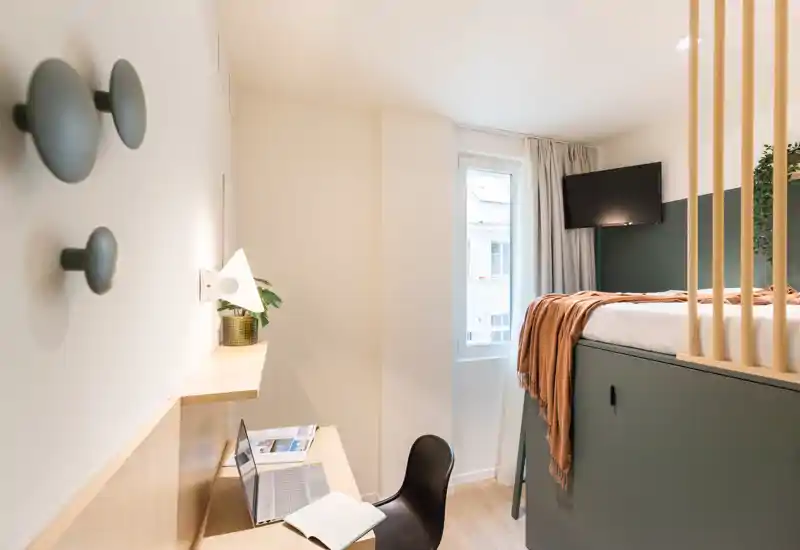 Camera luminosa e ben progettata con letto rialzato, coperta accogliente e scrivania vicino alla finestra — perfetta per un monolocale in affitto.
