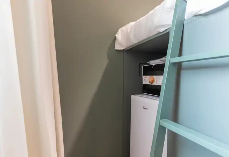 Angolo camera compatto con una cuccetta superiore visibile, scala e piccolo frigorifero sotto; stile pulito e minimal con toni neutri morbidi.