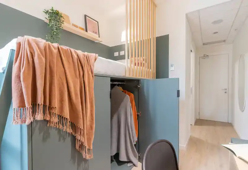 Camera singola contemporanea con letto rialzato e armadio integrato, tessuti caldi e illuminazione luminosa e ariosa per uno spazio compatto e accogliente.