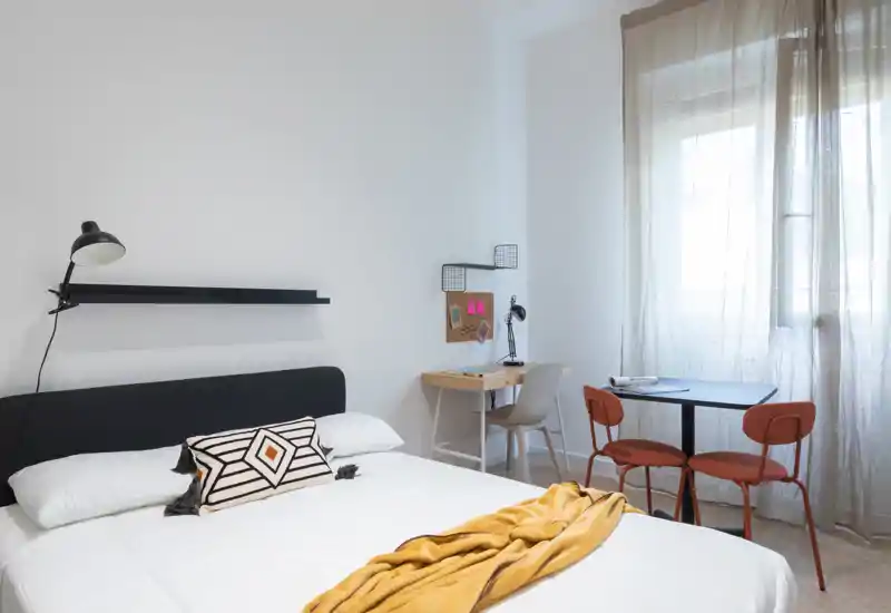 Bright, modern bedroom with a neatly made double bed, decorative cushion and a mustard throw. Natural light fills the room and a small desk and table with two chairs sit by the window, ideal for work or dining.