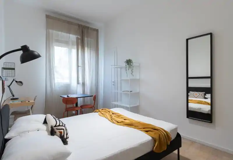 Bright, modern bedroom with a comfortable double bed, decorative cushions and a mustard throw. Natural light fills the room from a large window; minimal, stylish furniture includes a small desk and chairs.