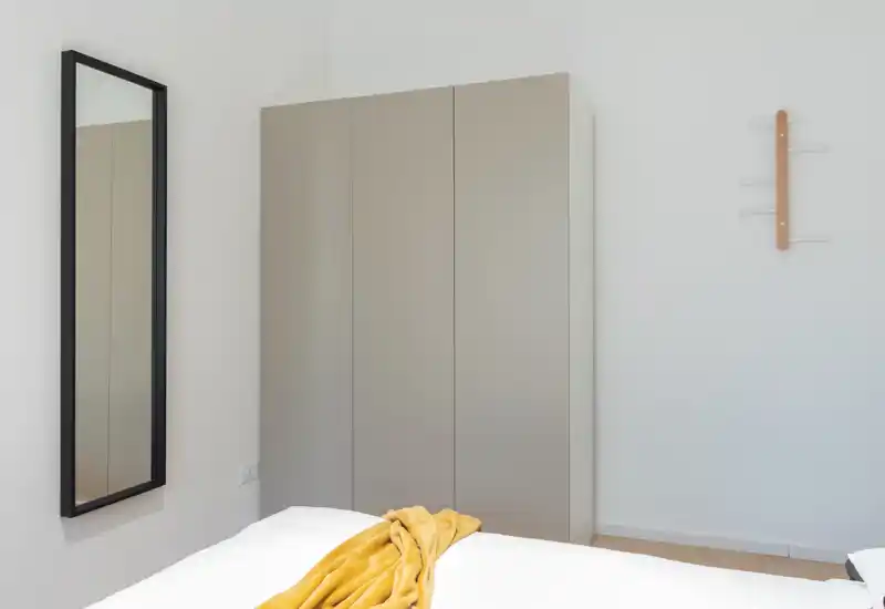 Bright, minimalist bedroom corner with a partial view of the bed, a tall wardrobe and a full-length mirror — simple, modern and well lit.