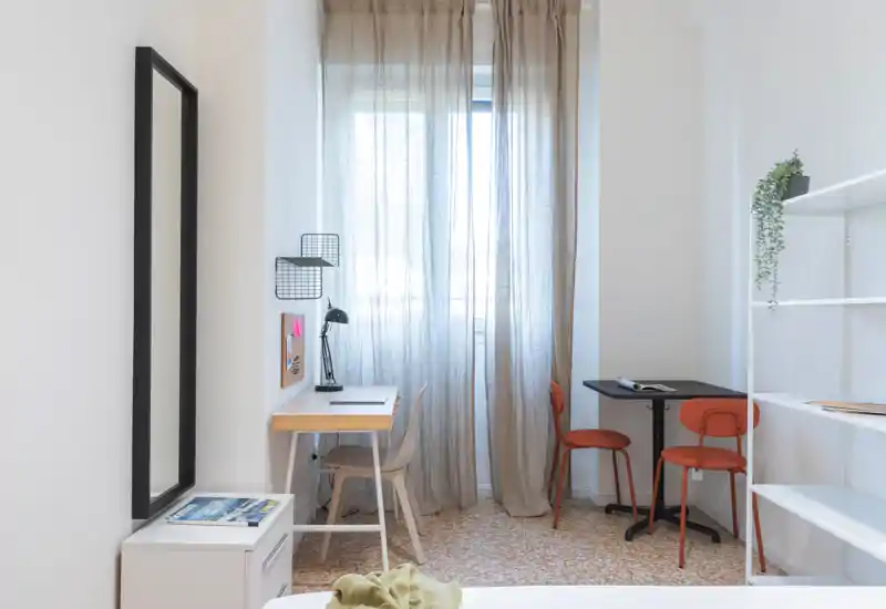 Bright, minimal bedroom with a bedside desk and small dining table by a sheer-curtained window; clean lines and warm accents create a calm, functional space.
