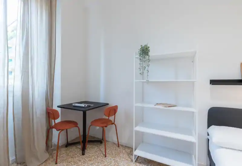 Bright, minimal bedroom corner with a small table and two chairs, a white shelving unit and the edge of a bed visible; natural light filters through sheer curtains.