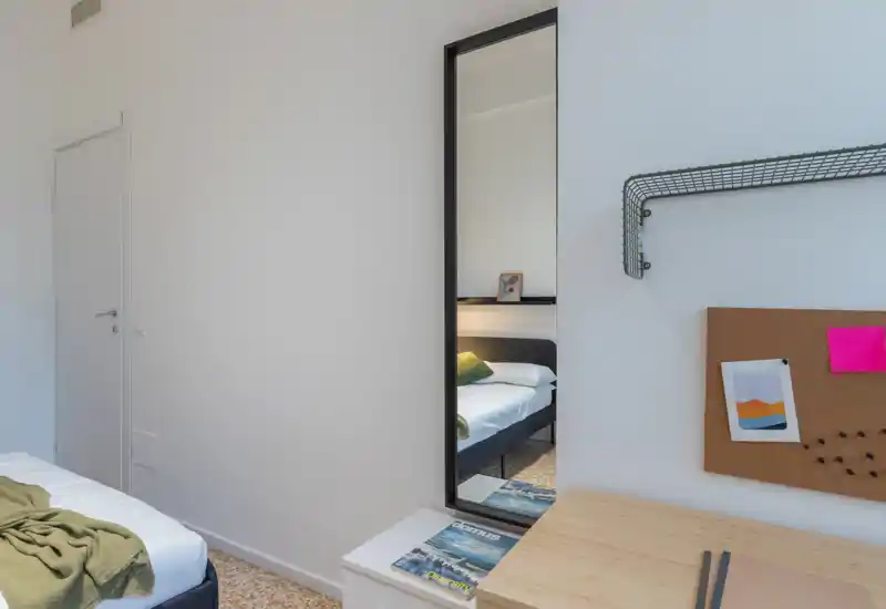 Compact, modern bedroom with a single bed visible directly and also reflected in a tall mirror; light, neutral walls and a small desk create a bright, functional space.