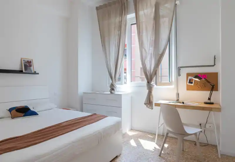 Bright, minimal bedroom with a double bed, bedside shelf and a small desk by the window creating a clean, functional space ideal for short-term rentals.