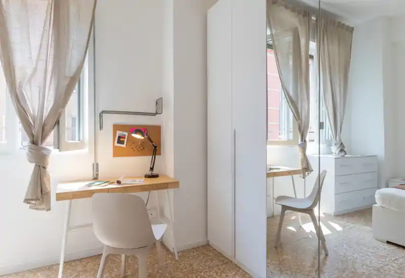 Bright, modern bedroom with a visible bed, desk by the window, and mirrored wardrobe that adds depth. Natural light and neutral textiles make the room feel airy and inviting.