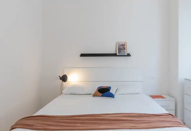 Bright, minimalist bedroom with a neatly made double bed, decorative throw and pillow, and a bedside lamp creating a warm accent.