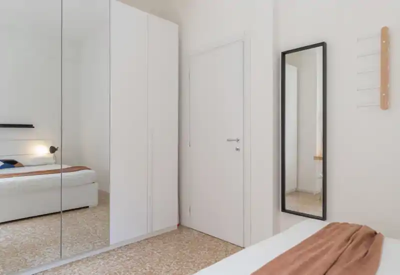 Bright, minimalist bedroom with a low-profile bed, mirrored wardrobe and warm throw. The room feels clean and airy with natural light and simple decor.
