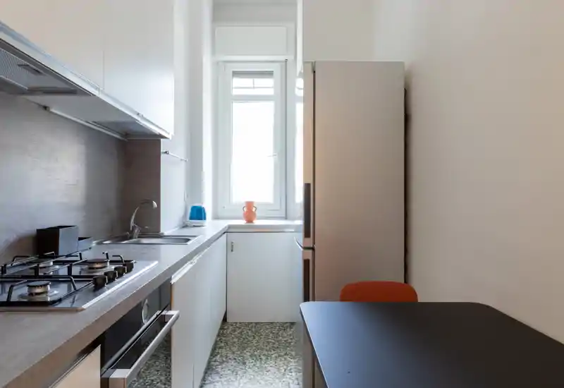 Compact modern kitchen with gas hob, built-in oven and a refrigerator, bright natural light from the window and a small dining surface—clean and functional layout ideal for a small apartment.