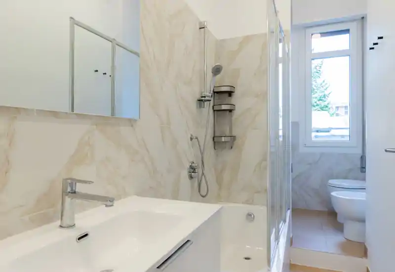 Modern bathroom with a bathtub and wall-mounted showerhead, white sink and bidet, marble-look wall tiles and a window providing natural light.