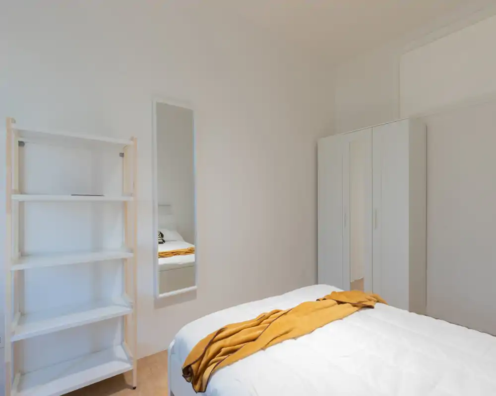 Minimal, bright bedroom with a white bed dressed in a quilt and a mustard throw; wardrobe and full-length mirror add practical storage and depth.