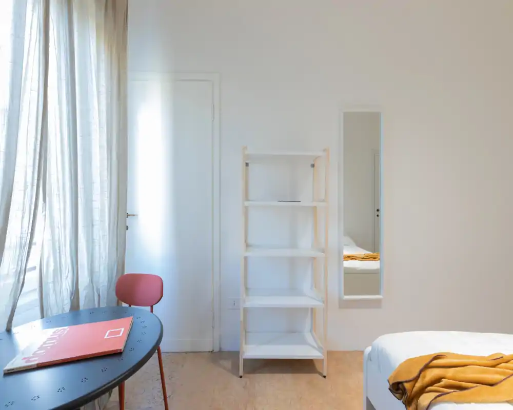 Minimal, bright bedroom corner with a partially visible bed dressed in white and a warm mustard throw; a shelving unit, slim mirror and a small table by a sheer-curtained window complete the scene.